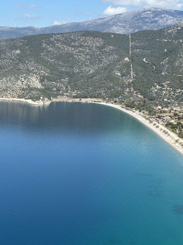 Πανοραμική άποψη από ψηλά της παραλίας Ψάθας, με γαλάζια θάλασσα, καμπύλη ακτογραμμή και βουνό στο φόντο